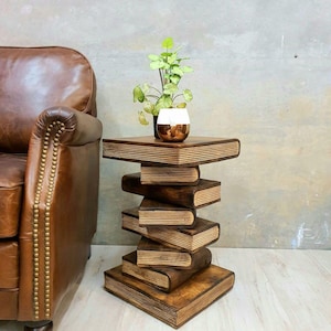 May include: A side table constructed from a stack of dark brown wooden books with visible page edges. A small green potted plant in a white and metallic gold ceramic pot sits on the top surface of the table, which is positioned next to a brown leather chair.