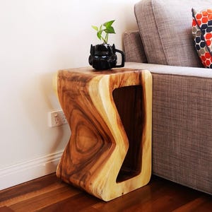 May include: A wooden side table with a unique, asymmetrical design. The table has a natural wood finish and a cutout in the side. A small potted plant sits on top of the table.