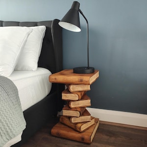May include: A side table constructed from a stack of six wooden books with a dark brown finish. A black desk lamp with a conical shade and a flexible neck is positioned on the top surface of the table, which is placed next to a bed.