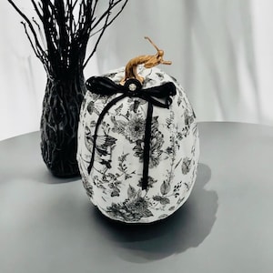May include: A white pumpkin decorated with a black and white floral and bird pattern. The pumpkin features a metallic gold stem and is adorned with a black ribbon tied in a bow at the top, centred with a small clear rhinestone embellishment.