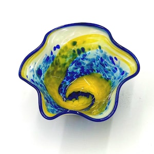 May include: A decorative glass bowl with a wavy rim. The bowl features a vibrant design of yellow and blue swirls and speckles, with a dark blue rim. The colors create a dynamic and artistic appearance, suitable for home decor.