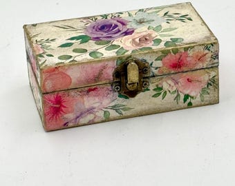 Può includere: Una scatola rettangolare in legno con un design floreale. La scatola presenta uno sfondo color crema con fiori in stile acquerello nei toni del rosa, viola e verde. Ha una chiusura con fermaglio in metallo.