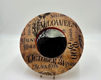 Op de afbeelding: Ronde decoratieve Halloween-spiegel met een zwart interieur en een rode rand. De buitenste ring heeft een vintage papieren ontwerp met Halloween-woorden en -zinnen, waaronder "October 31" en "Black Cats".