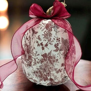 Puede incluir: Una calabaza decorativa con base blanca y un detallado patrón floral tipo toile en color rojo oscuro. La calabaza tiene un tallo de color dorado metálico y está adornada con un gran lazo de cinta transparente en color rojo oscuro.