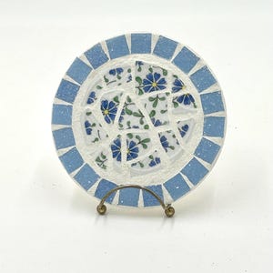 Puede incluir: Un posavasos de mosaico azul y blanco con un patrón floral. El posavasos está en un soporte de metal dorado.