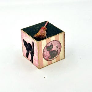 Könnte beinhalten: Ein dekorativer Holzquader mit schwarzer Oberseite und rosa-weiß gestreiften Seiten. Der Quader zeigt eine schwarze Katzen-Silhouette und eine Katzen-Illustration in einem ovalen Rahmen. Ein Miniatur-Besen liegt oben auf.
