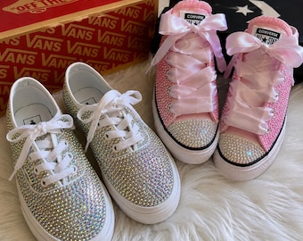 May include: Two pairs of bedazzled sneakers. One pair is silver, the other is pink. Both pairs have white soles and laces. The pink sneakers have large pink ribbon laces. A red Vans box is in the background.