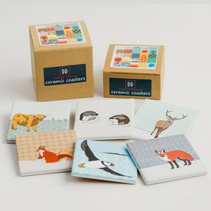 May include: Set of six ceramic coasters featuring various animal illustrations. The coasters are packaged in two brown boxes with the text "Dibujo Design ceramic coasters". The illustrations include a Highland cow, two otters, a red deer, a fox, and a puffin.