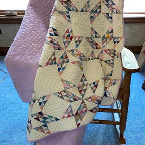 May include: A quilt featuring a star pattern composed of various multi-colored fabric scraps on a cream background. The quilt has a solid light purple backing and border, with a quilted texture throughout, draped over a wooden rocking chair.