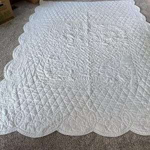May include: A solid white quilt featuring a diamond-stitched pattern across the center and a scalloped edge border. The fabric displays an embossed floral design throughout, with a central motif of a basket filled with flowers and foliage.