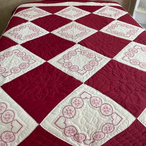 May include: A quilted bedspread featuring a diamond pattern in white and burgundy. The white diamonds are embroidered with a pink floral design. The burgundy diamonds have a quilted floral pattern.
