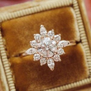 May include: A gold ring featuring a central clear stone encircled by smaller, petal-shaped gems. The ring is presented in a velvet box. The design is reminiscent of a flower or starburst.