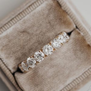 Peut inclure: Une bague en or ornée de cinq pierres précieuses rondes et transparentes, disposées en ligne. La bague est présentée dans un écrin rectangulaire ouvert, doublé de velours marron clair. L'écrin met en valeur le bijou.