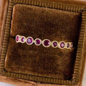 May include: A gold ring with a series of round, deep red gemstones set in a row. The ring is displayed inside a dark brown velvet ring box. The gemstones are evenly spaced and surrounded by a delicate, beaded design.