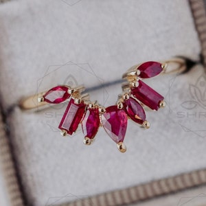 May include: A gold ring featuring a fan-shaped arrangement of bright red gemstones. The ring is presented in a velvet-lined jewellery box, highlighting the ring's intricate design and colour contrast. The gemstones are different shapes, enhancing the ring's unique appeal.