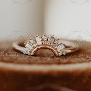 May include: A gold-colored ring with a half-circle design, adorned with multiple small, clear gemstones. The ring is set on a brown velvet surface, showcasing its intricate details and elegant style.