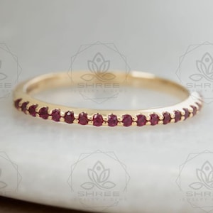 May include: A gold ring featuring a line of small, round, red gemstones. The ring is displayed on a white surface. The gemstones are evenly spaced around the band.