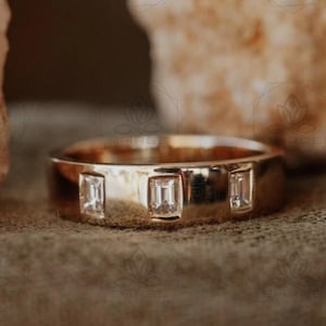 May include: A gold band ring featuring three rectangular, clear gemstones. The ring is presented on a textured, neutral-toned surface, with blurred, light-coloured rocks in the background. The ring's design is simple and elegant, suitable for everyday wear.