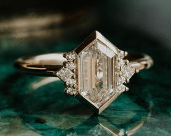 May include: A gold ring with a large, clear, geometric-cut center stone. The ring features smaller accent stones on either side of the center stone. The ring is set against a green and gold background.