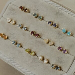 May include: A collection of stud earrings with various colored gemstones, including red, blue, green, and white, set in gold. The earrings are arranged in pairs inside a gray jewelry case.