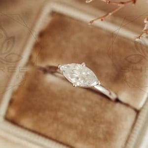 May include: A marquise cut diamond engagement ring set in a silver band, displayed in a beige velvet ring box.