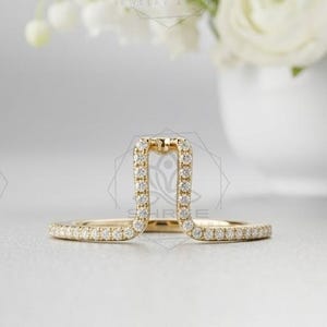 Peut inclure: Une bague en or sertie de petits diamants étincelants. La bague a un design unique avec une forme rectangulaire verticale au centre. La bague est présentée sur une surface blanche avec un bouquet de fleurs blanches en arrière-plan.