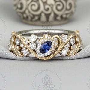 May include: A two-tone metal ring featuring a central marquise-cut blue sapphire surrounded by small round white stones. The band is designed with intricate gold-toned leaf motifs and silver-toned metal accents, creating a detailed botanical pattern.