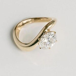 May include: A gold ring featuring a single round brilliant-cut diamond set in a bypass-style band. The polished yellow gold band curves gracefully around the center stone, which is secured by multiple prongs, creating a fluid and continuous silhouette.