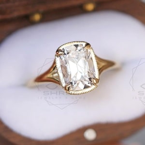 May include: A gold engagement ring with a rectangular, clear gemstone is displayed inside a wooden ring box. The ring has a delicate, detailed design and is set against a white interior.
