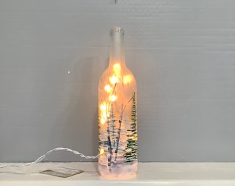 May include: A frosted glass bottle with a winter scene painted on it. The bottle is illuminated by warm white fairy lights inside. The painted scene depicts trees and a snowy landscape. The bottle is approximately 12 inches tall.