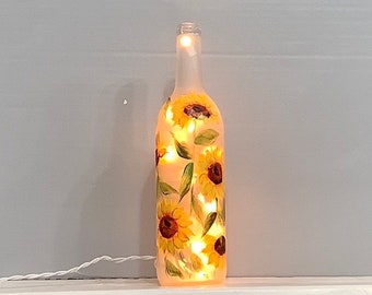 May include: A frosted glass wine bottle with a painted sunflower design and string lights inside. The bottle is lit up and the lights are visible through the frosted glass.