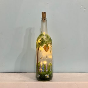 May include: A frosted glass wine bottle with a cork top, painted with a green tree, a yellow honeycomb, and purple flowers.  There are small yellow bees flying around the honeycomb.