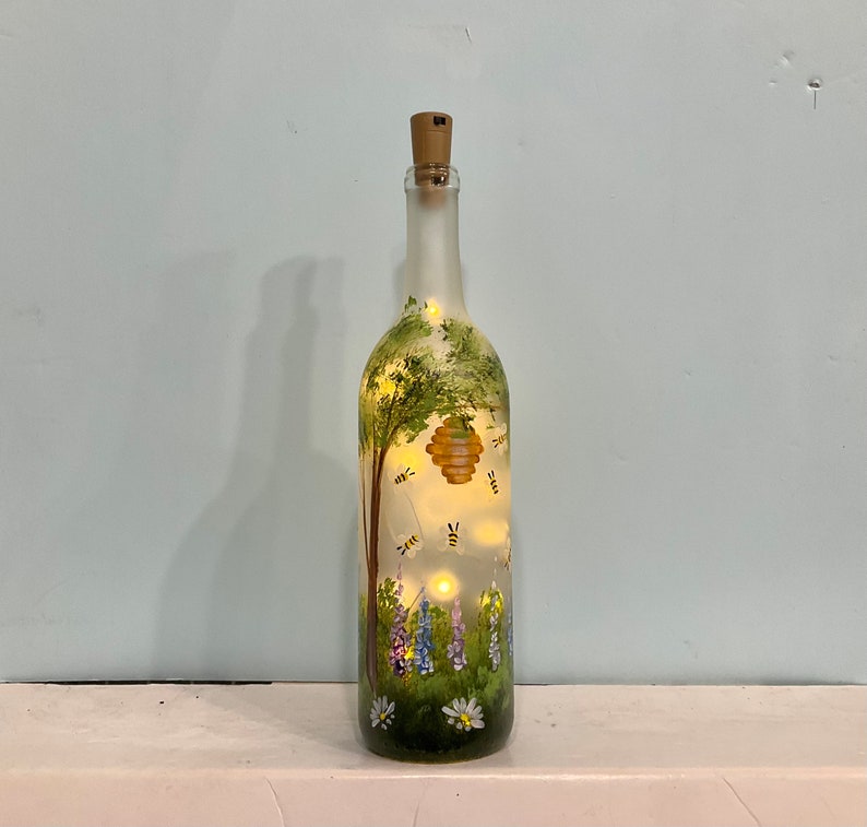 May include: A frosted glass wine bottle with a cork top, painted with a green tree, a yellow honeycomb, and purple flowers.  There are small yellow bees flying around the honeycomb.