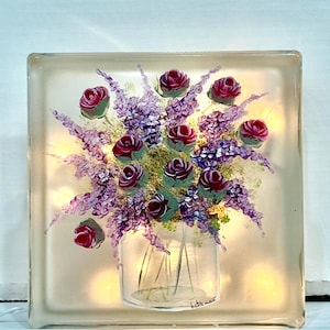 May include: A glass block with a painted image of a bouquet of purple and pink flowers in a clear glass vase. The flowers are painted in a realistic style and the vase has a simple design. The glass block is illuminated from behind, creating a warm glow.