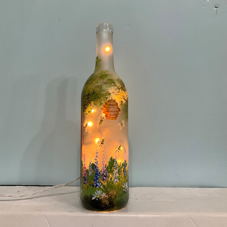 May include: A glass wine bottle with a painted design of a beehive, bees, and wildflowers. The bottle is lit from inside, creating a warm glow.