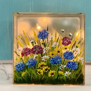 May include: A square glass block with a painted floral design. The artwork features blue, yellow, white, and burgundy flowers with green grass and stems. Small lights are visible inside the block, creating a warm glow. The block is lit by a string of lights.