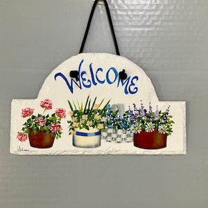 May include: A white slate sign with the word "Welcome" painted in blue. The sign is decorated with painted flowers in pots, including pink geraniums, blue and white flowers, and lavender.