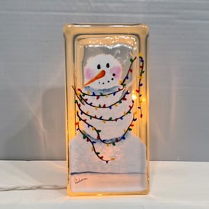 May include: A hand-painted glass block with a snowman design. The snowman is wearing a blue scarf and is surrounded by colorful Christmas lights. The block is lit from within, creating a warm glow.
