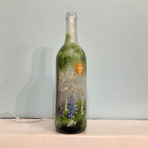 May include: A frosted glass wine bottle with a painted scene of a beehive, bees, flowers, and a tree. The bottle is lit from inside, creating a warm glow.