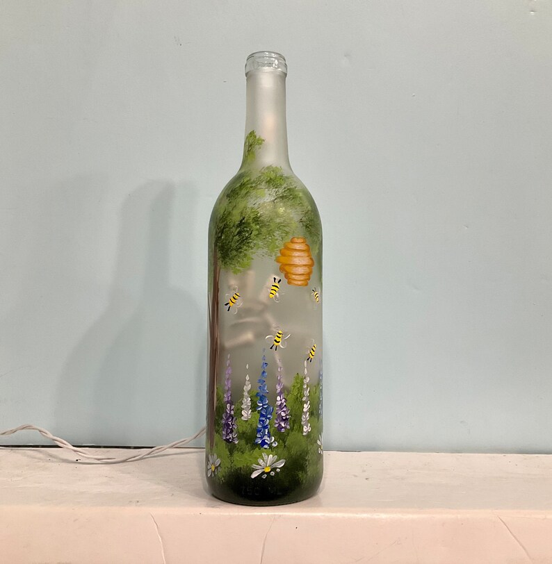 May include: A frosted glass wine bottle with a painted scene of a beehive, bees, flowers, and a tree. The bottle is lit from inside, creating a warm glow.