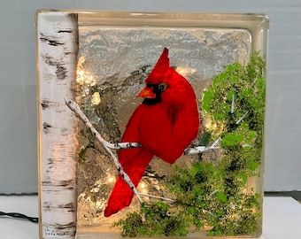 May include: A red cardinal bird perched on a branch with green foliage, painted on a clear glass block with a white birch tree in the background. The block is illuminated with white lights.