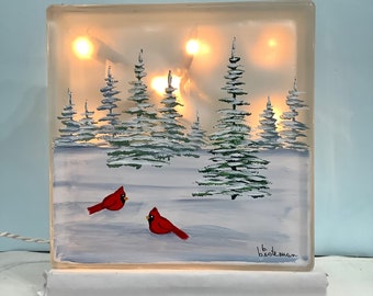 May include: A lighted glass block featuring a winter scene with two red cardinals in a snowy landscape. The artwork includes snow-covered evergreen trees and a soft, glowing light from within. The block is on a white base.