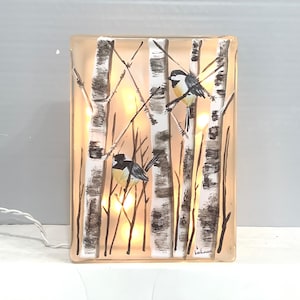 May include: A glass block with a painted scene of birch trees and two birds. The block is illuminated with warm white LED lights. The signature "backman" is visible in the bottom right corner.