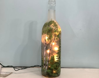 May include: A frosted glass wine bottle with a painted scene of birch trees and a forest inside. The bottle is lit from within with warm white lights.