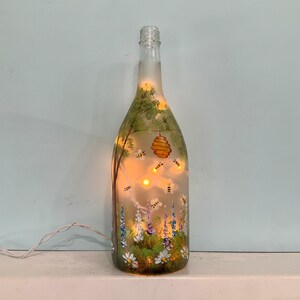May include: A glass bottle with a frosted finish, hand-painted with a scene of a beehive, bees, flowers, and trees. The bottle is lit from inside, creating a warm glow.