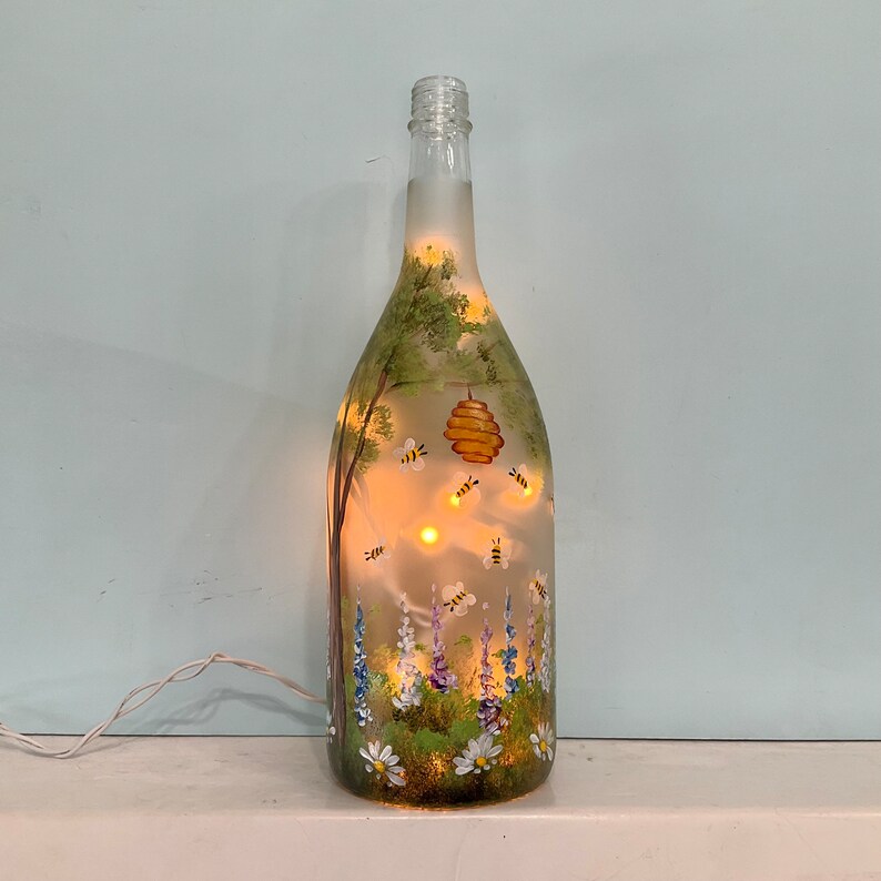 May include: A glass bottle with a frosted finish, hand-painted with a scene of a beehive, bees, flowers, and trees. The bottle is lit from inside, creating a warm glow.