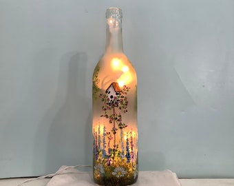 May include: A clear glass bottle with a frosted finish, painted with a whimsical scene of a birdhouse in a garden with flowers. The bottle is lit from within, creating a warm glow.