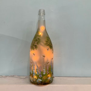 May include: A clear glass bottle with a floral and bee design painted on it. The bottle is lit from inside with white fairy lights.