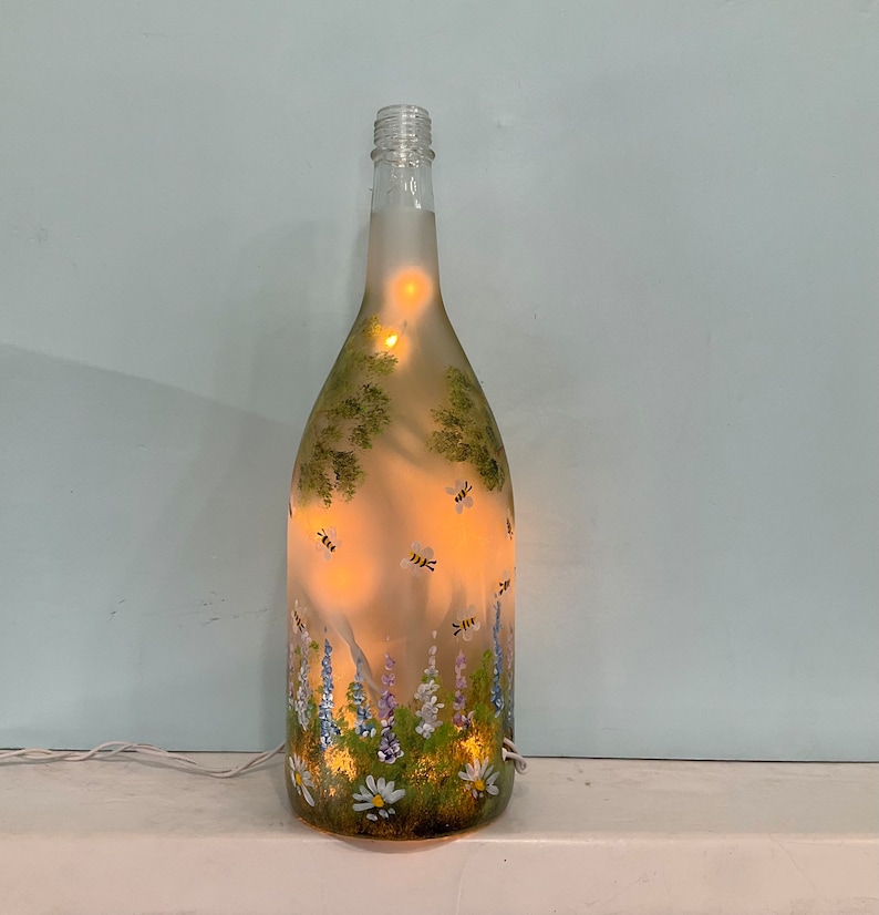 May include: A clear glass bottle with a floral and bee design painted on it. The bottle is lit from inside with white fairy lights.