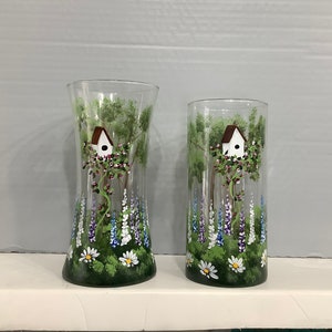 May include: Two clear glass vases with hand-painted floral designs. Each vase features a small birdhouse with a white roof and a green, white, and purple floral pattern.